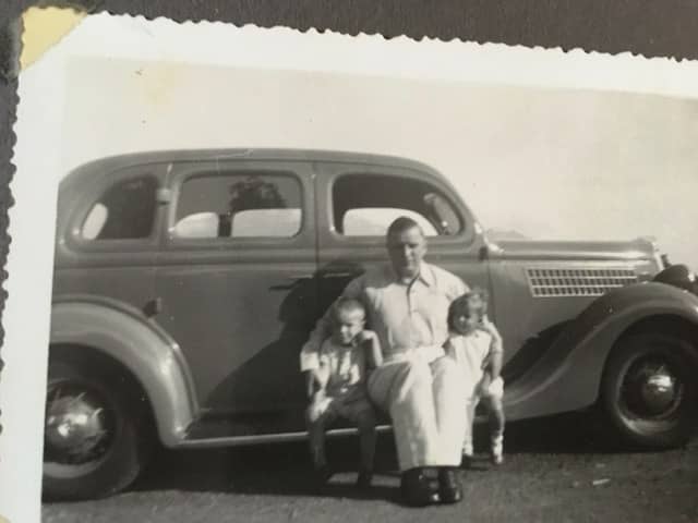 Zwart-witfoto. Een man zit met twee kinderen op de opstaprand van een auto. Een jongetje zich links naast hem, een meisje links, hij houdt zijn beide armen om hun schouders. Het jongetje steunt met zijn hoofd op zijn linkerhand en zijn linker ellenboog op de schoot van de man. Zowel de man als de kinderen dragen kleding in lichte kleuren.