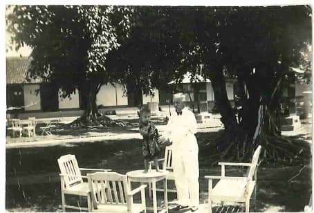 Zwart-witfoto. Een man staat tussen vier witte tuinstoelen naast een kleine, ronde tafel. Op het tafeltje staat een kleine jongen. De man draagt een wit pak met zwarte strik en strekt zijn armen uit naar het kind. De jongen draagt een traditioneel Indonesisch gewaad. In de achtergrond een grote boom met veel wortels boven de grond en de rest van de tuin: gras, planten, een schuurtje, een galerij.