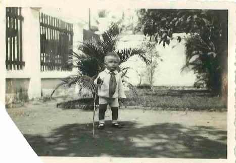 Zwart-witfoto. Een jongetje staat in een tuin. Hij draagt een pakje met een grote strik, hij houdt een stok in zijn rechterhand. Achter hem een varen en andere struiken, links een tuinhek.