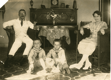 Zwart-witfoto. Groepsfoto van een gezin in een woonkamer. De man zit links in een rieten stoel, hij draagt een volledig wit pak met zwarte stropdas en een bril. De vrouw zit rechts in een rieten stoel, zij draagt een lichtgekleurde jurk. Tussen hen in zitten twee jongetje op de grond, zij dragen matrozen pakjes. Tegen de muur in de achtergrond staat een piano.