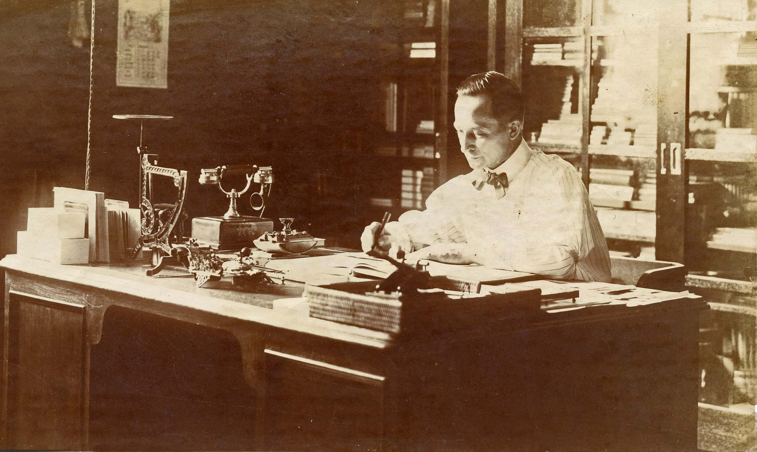 Zwart-witfoto. Een man zit achter een bureau te werken. Hij draagt een wit overhemd met zwarte strik. Op het bureau zijn meerdere attributen te zien, zoals een telefoon, een weegschaal, een inktdepper, veel papier in of naast bakjes. Achter de man een kast met glazen deuren vol met boeken en meer papieren.