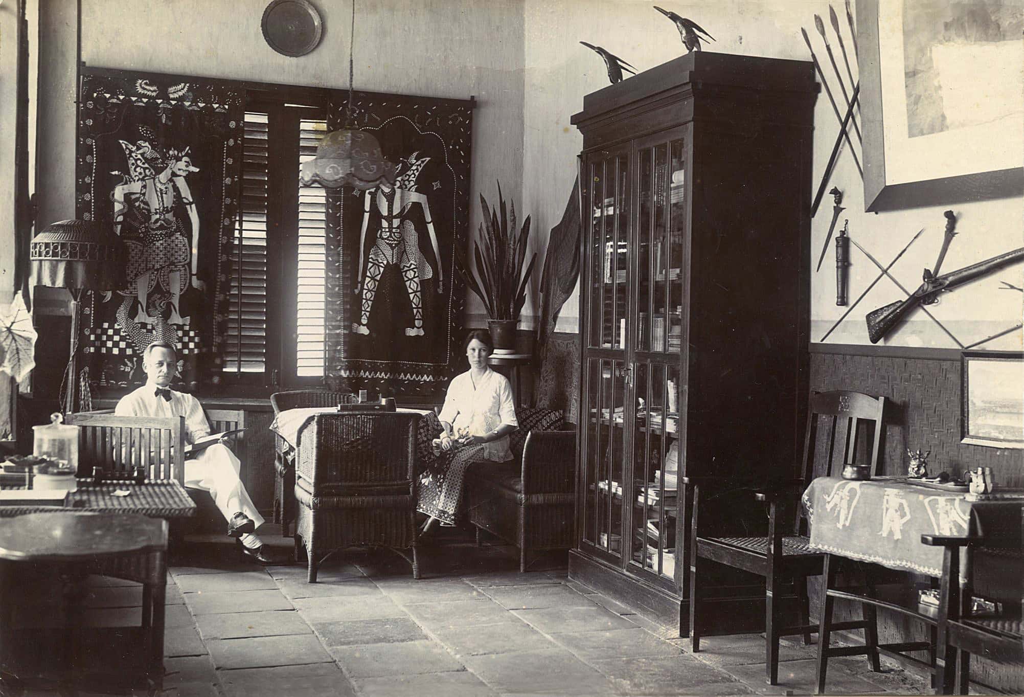 Zwart-witfoto. Een man en een vrouw zitten in een kamer. De man zit links in een houten stoel, de vrouw rechts van hem op een rieten bank. In het midden van de foto een grote houten kast met glazen deuren. In de kamer zijn meerdere tafels en stoelen. Aan de muur rechts hangen speren, een kris en een geweer. Voor de ramen achter de man en vrouw hangen gordijnen met Indonesische poppen erop.