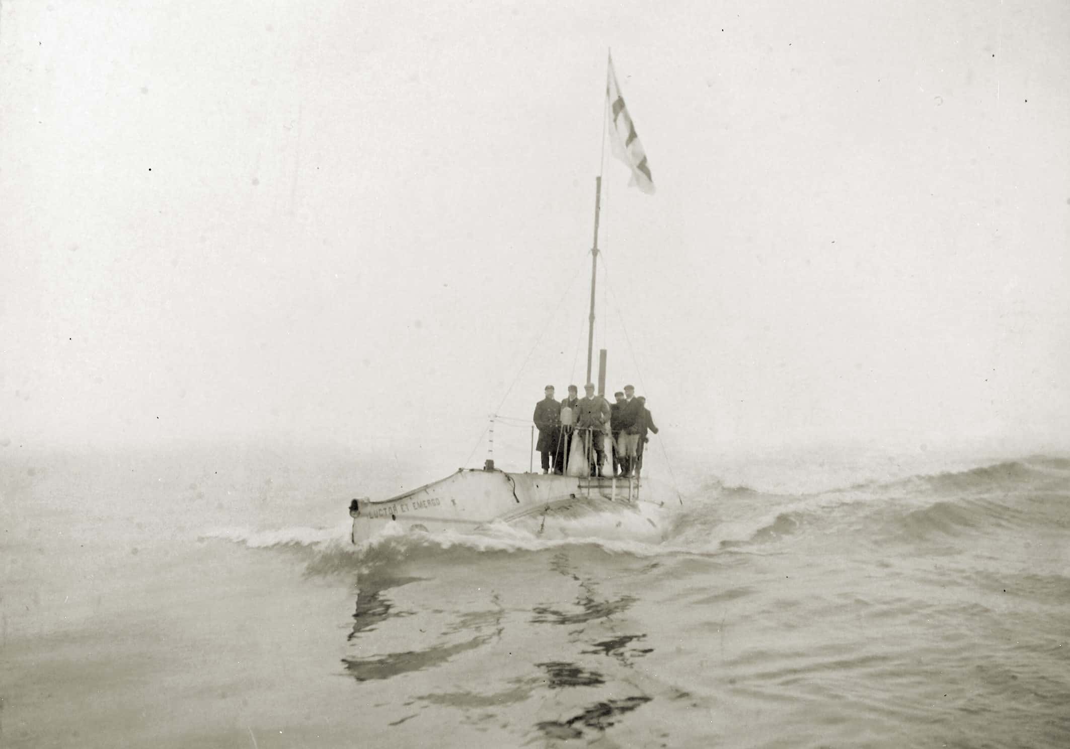 Zwart-witfoto. Een onderzeeboot in de zee. Nog net boven water. Op de bovenkant staat wat personeel. Op de boeg staat 'Luctor et emergo'.