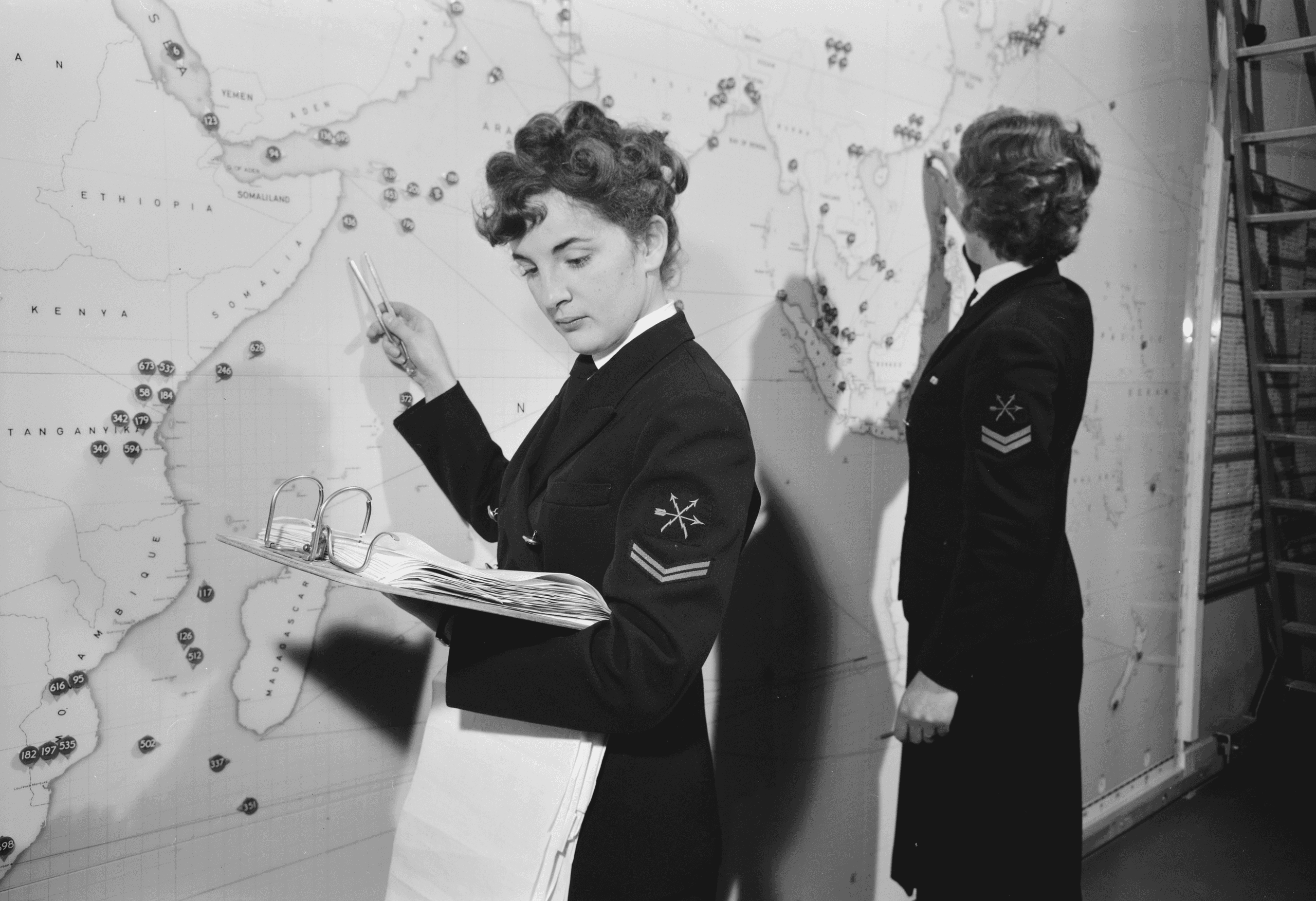 Zwart-witfoto. Twee vrouwen in militair uniform staan voor een kaart.