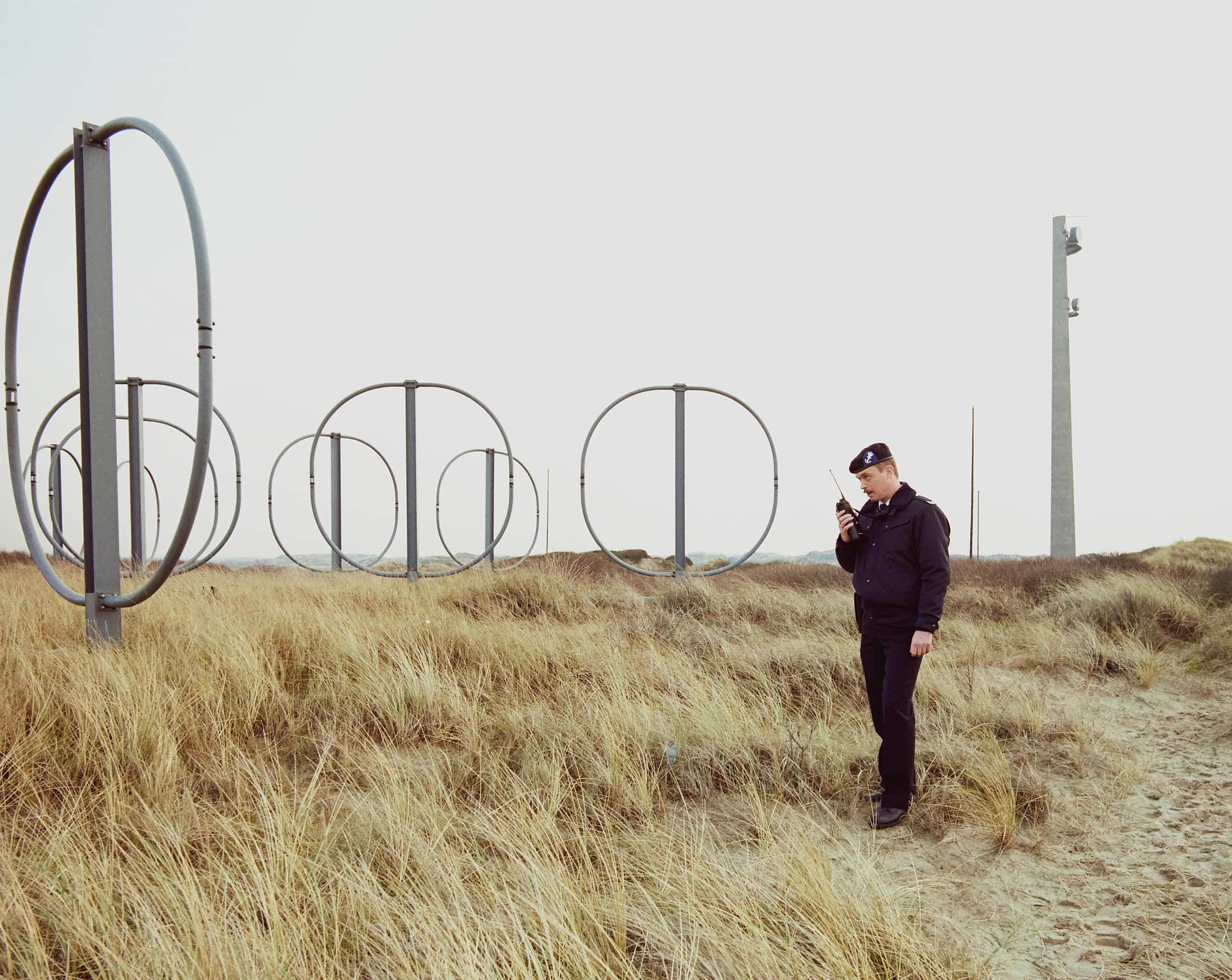 Kleurenfoto. Man in uniform spreekt in zijn portofoon. Hij staat in de duinen en is omringd door radio-masten.