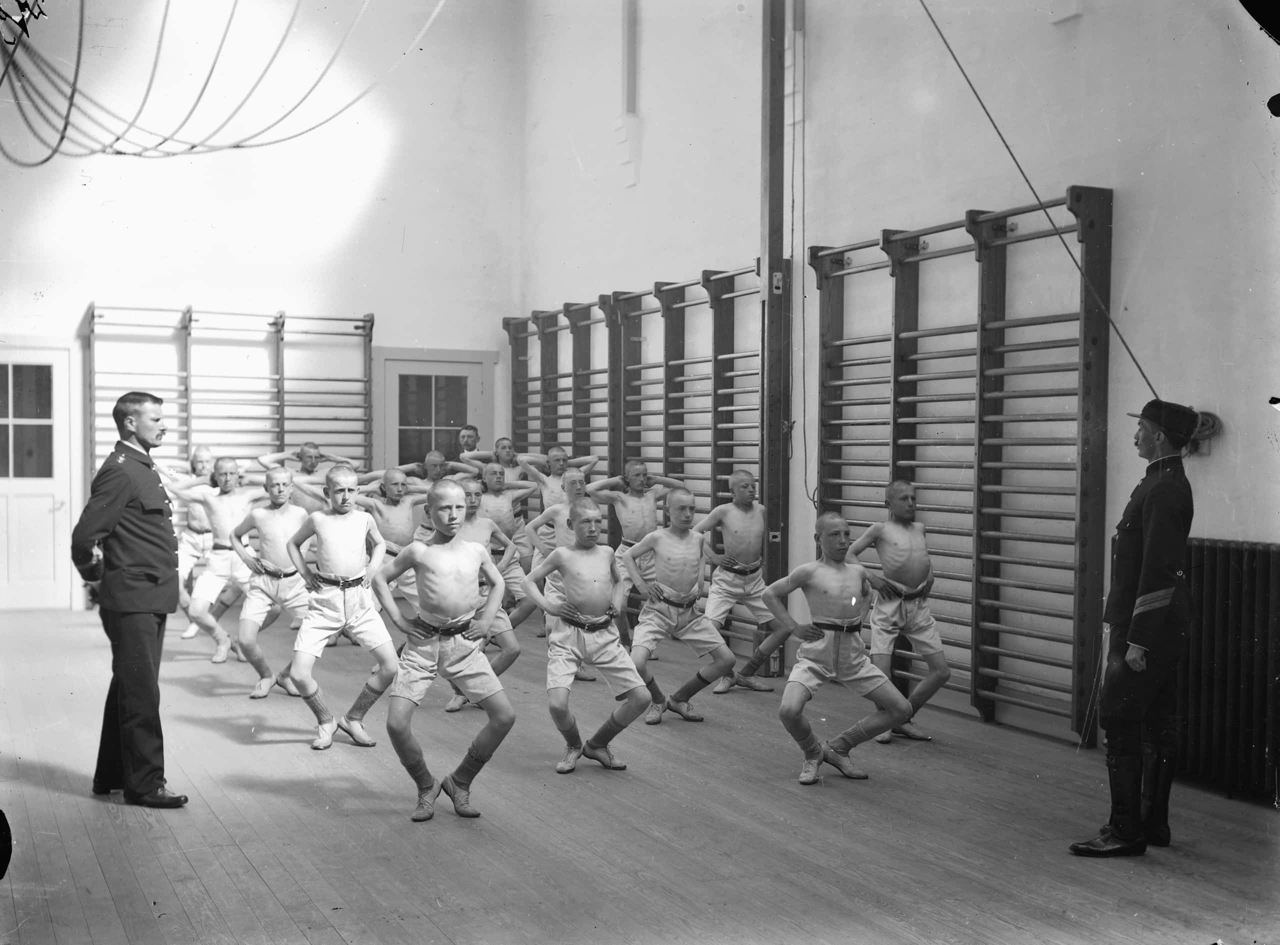 Zwart-witfoto. Een groep jonge jongens is te zien die gymnastische houdingen aannemen. Naast hen staan twee mannen in uniform te kijken.