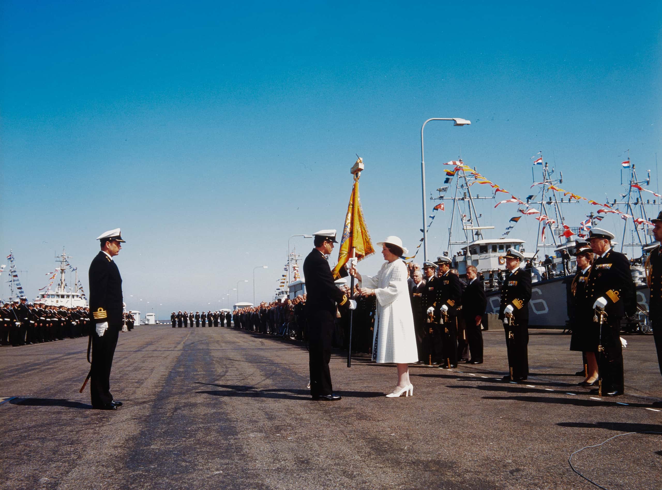 Kleurenfoto. Rijen aan marine-personeel is te zien. In het midden is het leeg. Koningin Beatrix, in het wit gekleed, draagt een vaandel over aan een marineofficier.