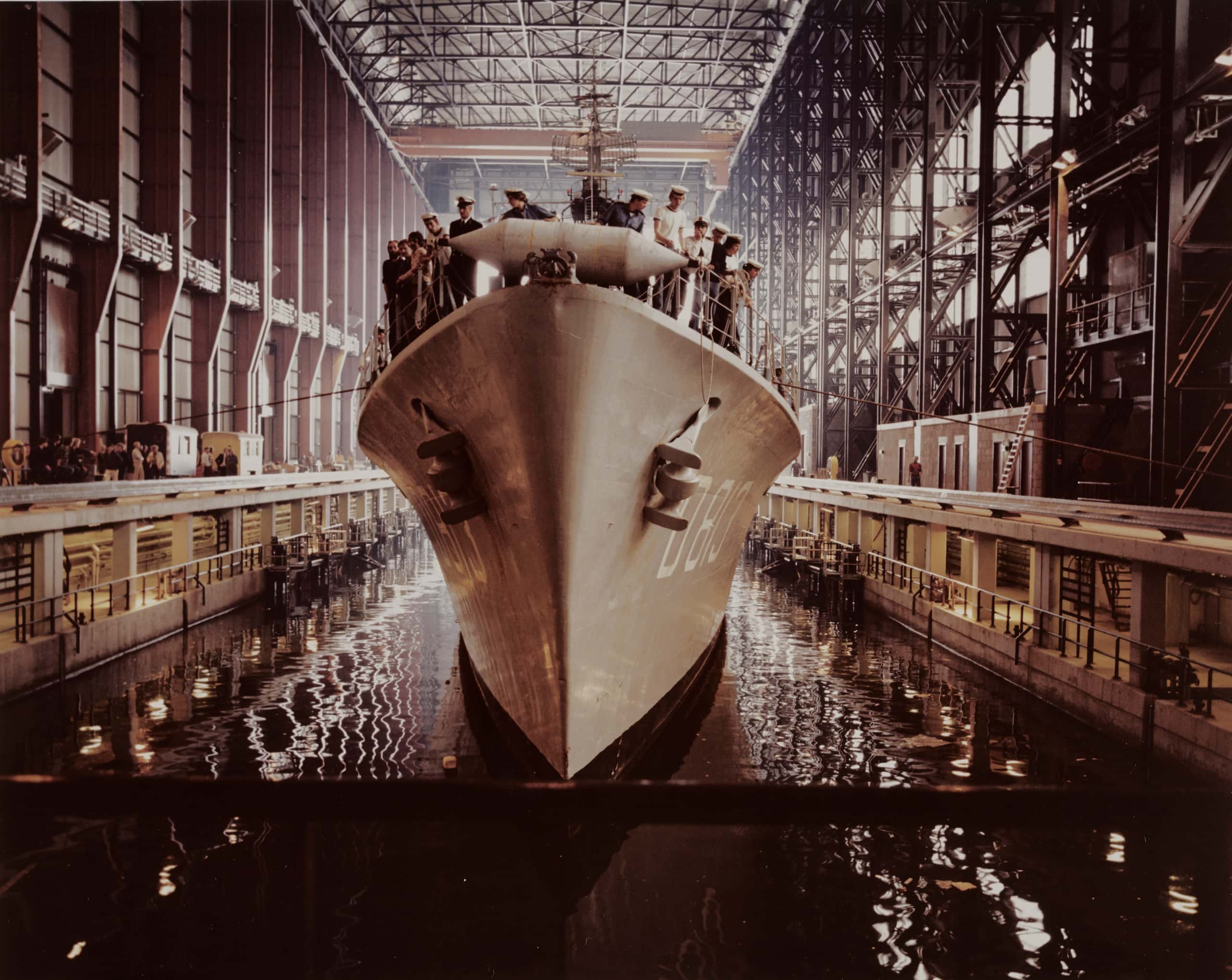 Kleurenfoto. Vooraanzicht van een marineschip. Het schip ligt in de scheepswerf. Op het dek staat marinepersoneel.