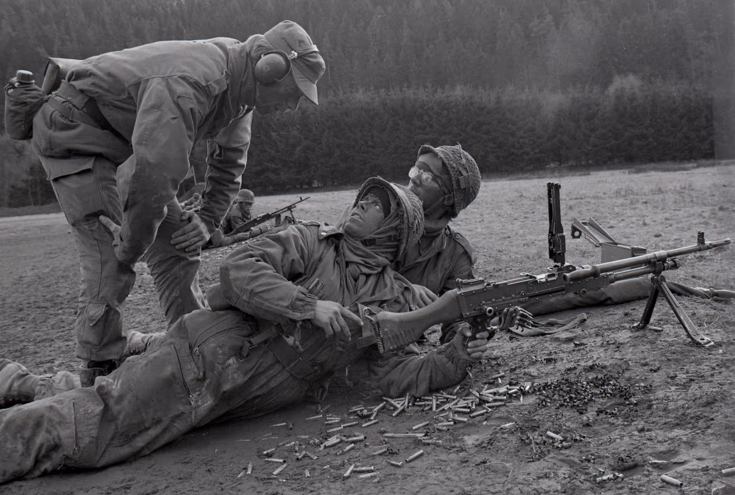 Zwart-wit foto. Er zijn drie militairen te zien, in uitrusting. Een militair staat voorovergebogen, de andere twee militairen liggen op de grond. Een van de militairen heeft een wapen.