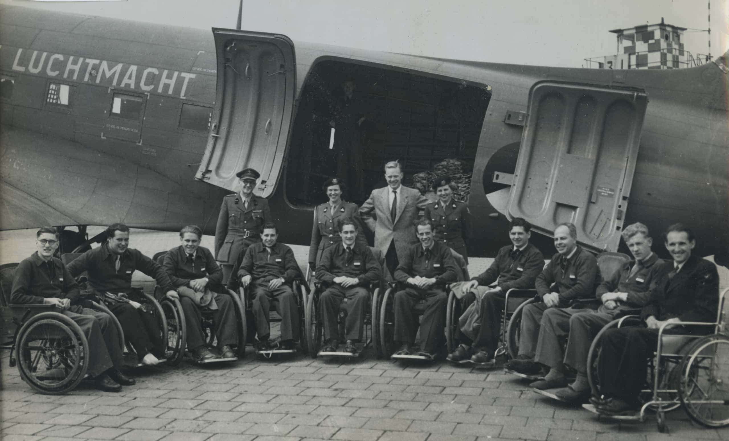 Zwart-witfoto. De zijkant van een vliegtuig met 'Luchtmacht' erop is te zien. Voor dit vliegtuig is een groep mensen te zien die poseren voor de foto. Tien personen zitten in een rolstoel. Daarachter staan vier personen: twee vrouwelijke militairen, een mannelijke militair en een burger.