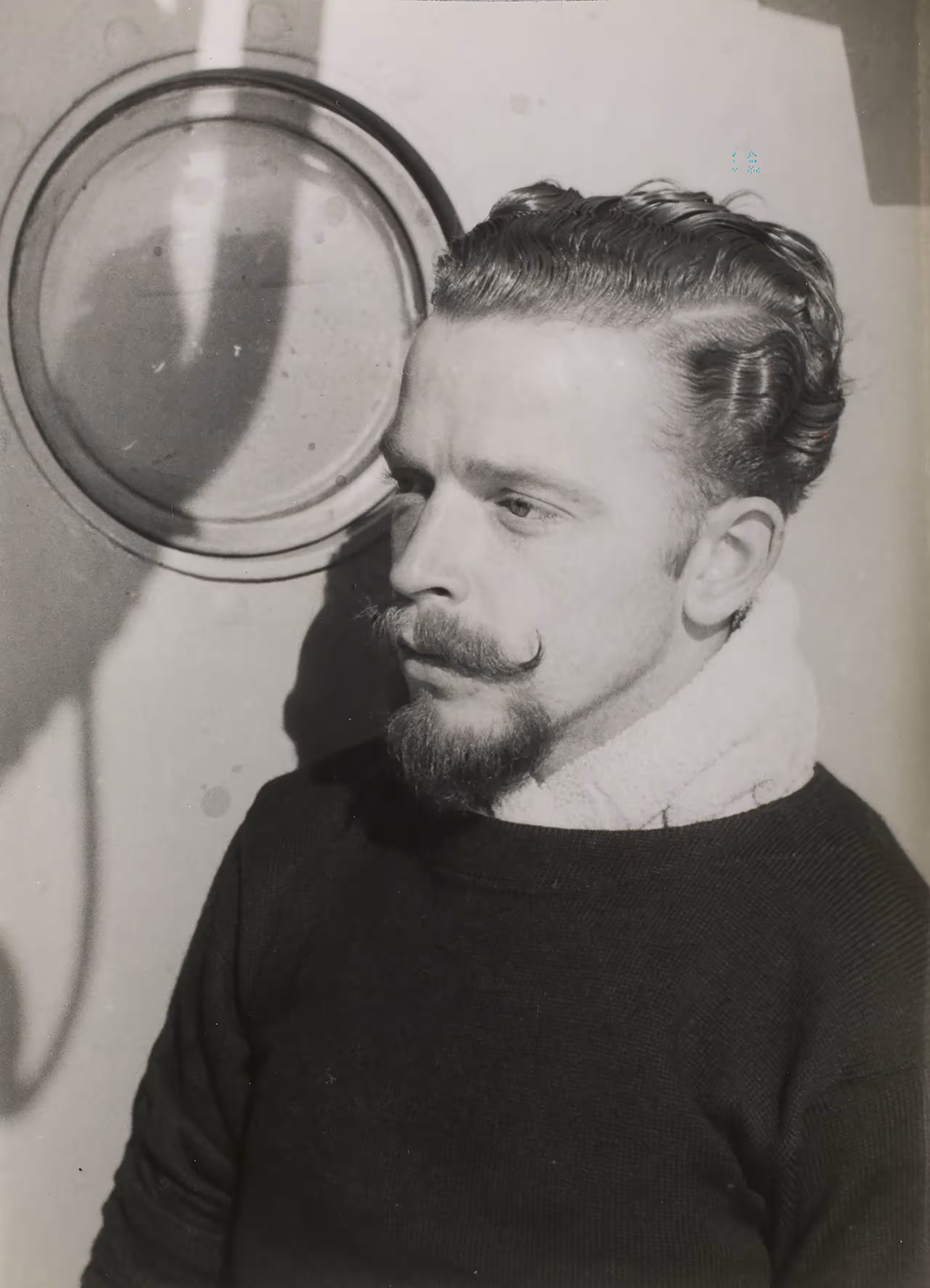 Zwart-witfoto. Een man kijkt naar links buiten de camera, zijn haar is gekamd met een lichte slag, zijn snor is in een punt naar boven gekruld, hij heeft een sik. Hij draagt een donkere trui met een handdoek om zijn nek, die in de trui is gestopt. Aan de muur achter hem een dichte patrijspoort.