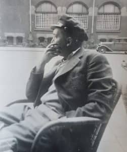 Zwart-witfoto. Man zit en kijkt naar de zijkant. Hij heeft een pet op en lange jas aan.