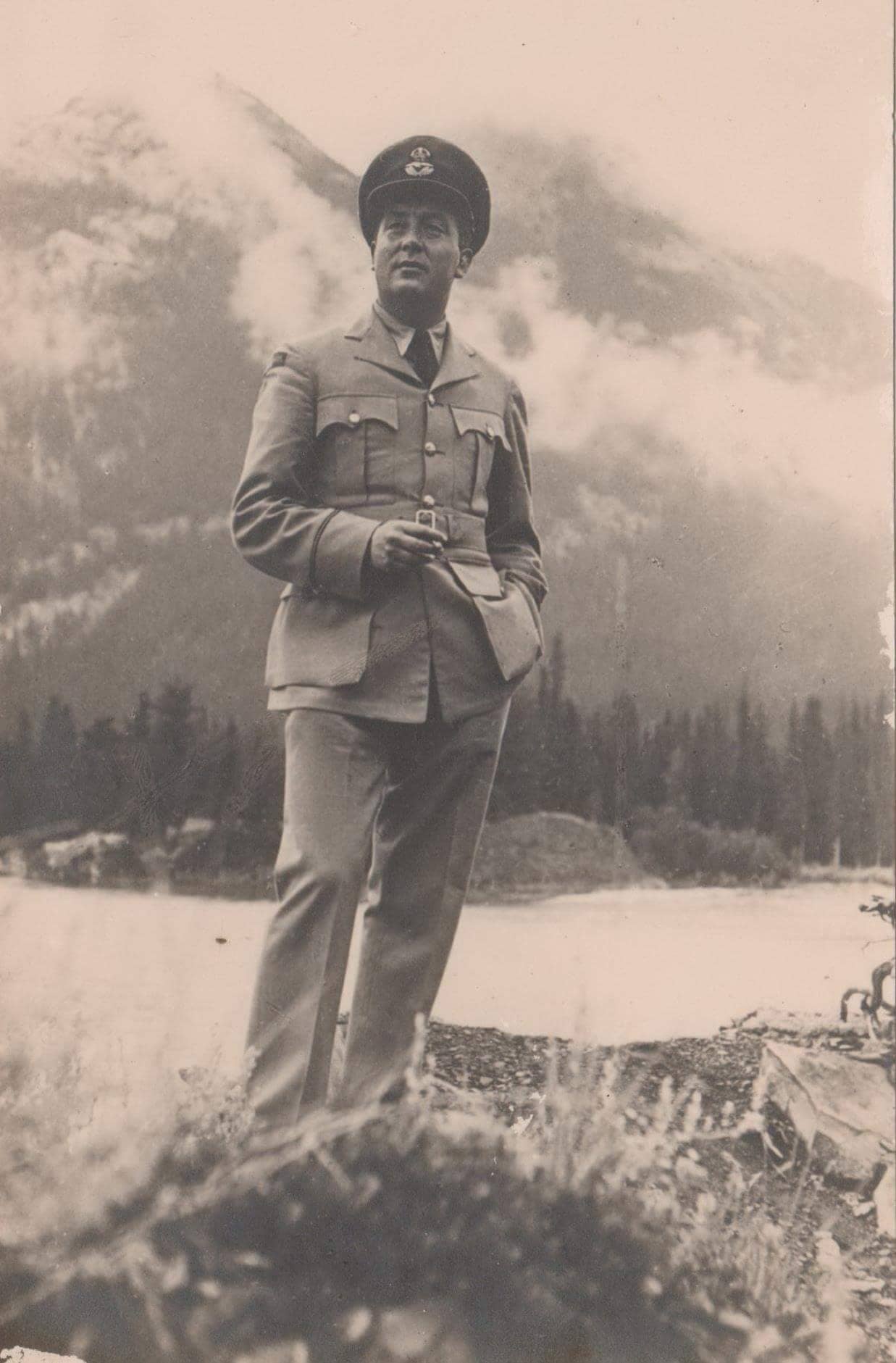 Zwart-witfoto, sepia. Een man in militair uniform poseert op een verhoging voor een rivier. Op de achtergrond zijn hoge bergen te zien.