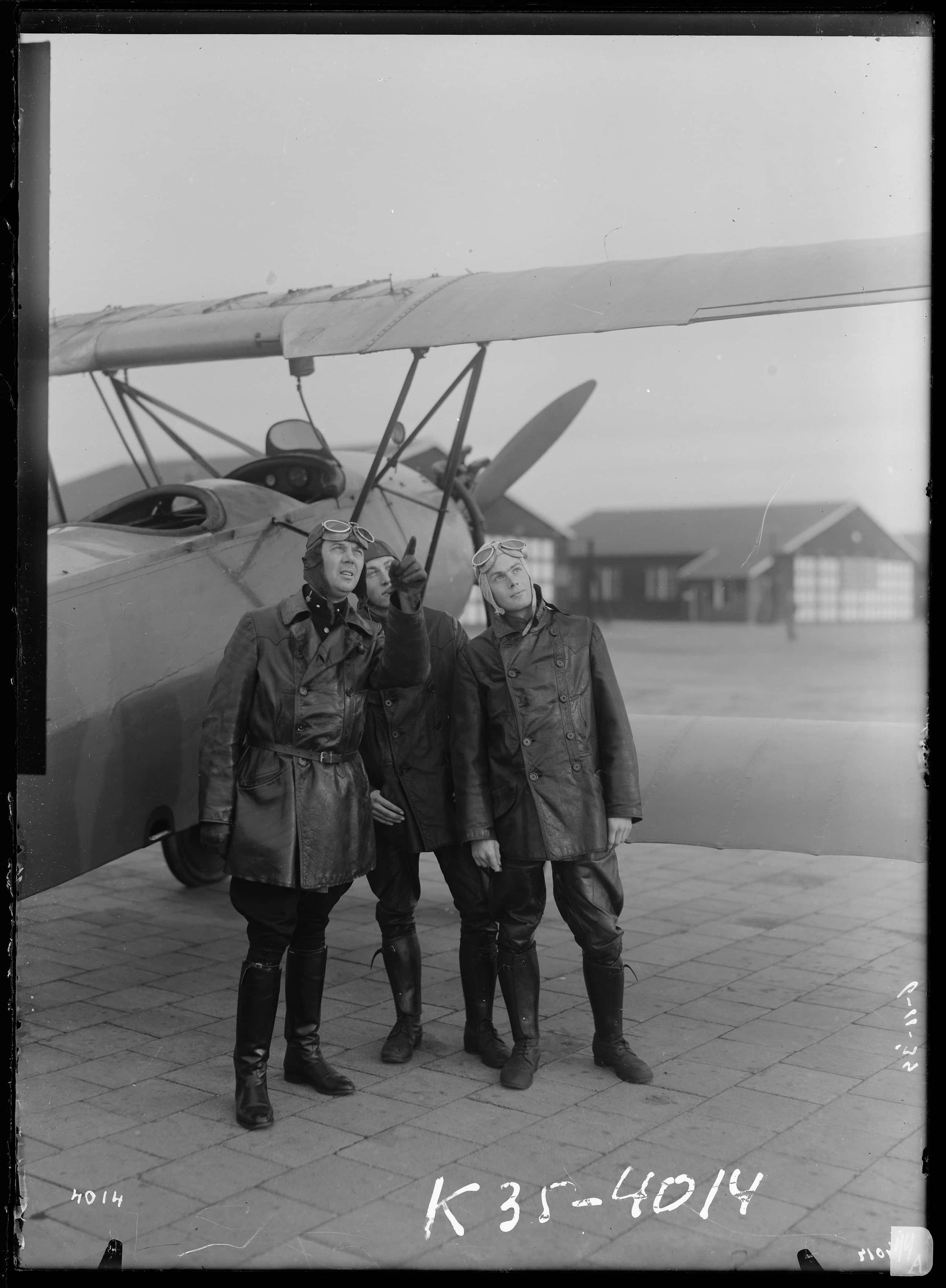 Zwart-witfoto. Drie mannen in militair vliegeruniform voor een vliegtuig kijken naar iets in de lucht.