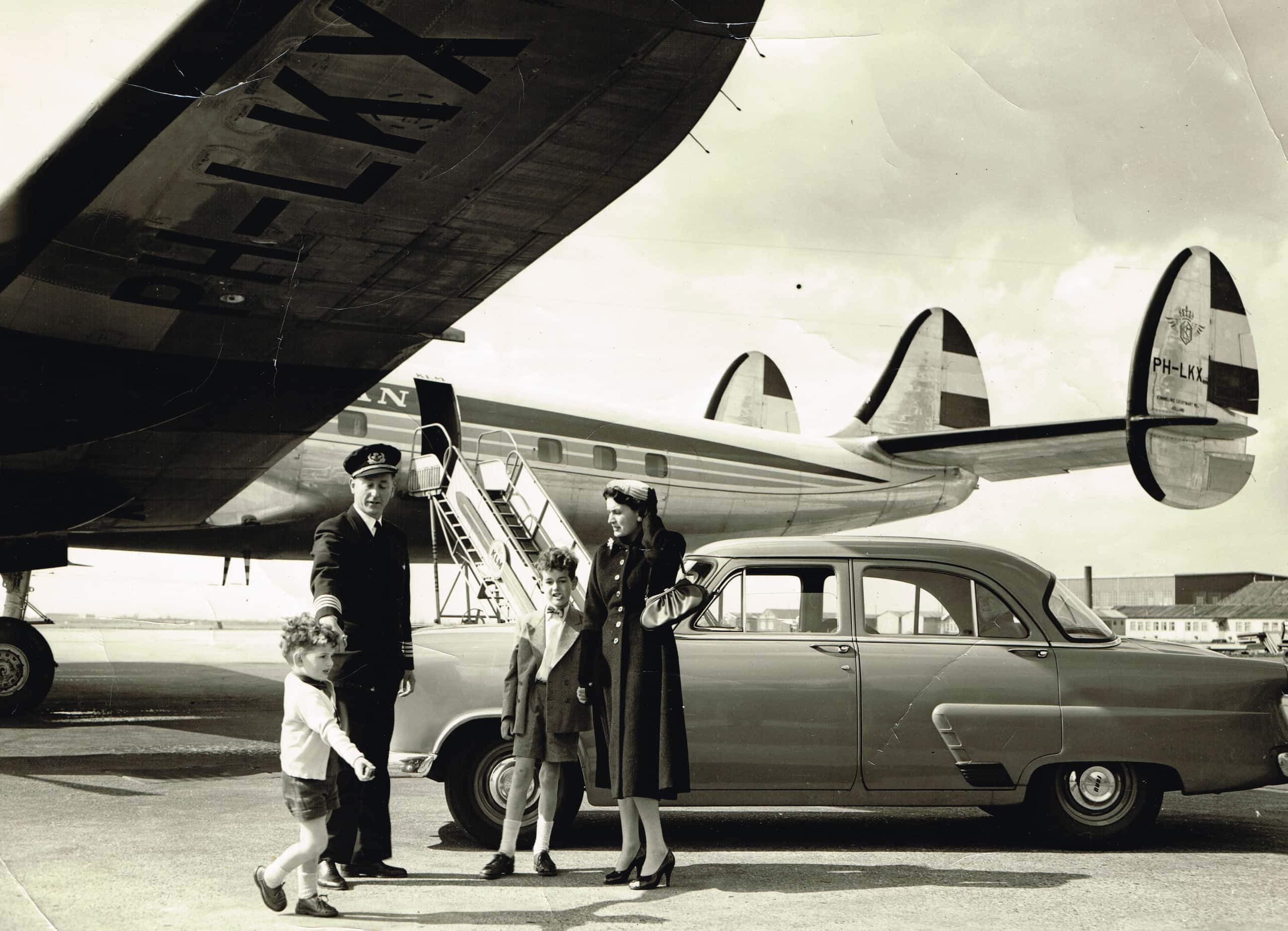Zwart-witfoto. Een gezin staat naast een auto. Daarachter staat een groot vliegtuig.