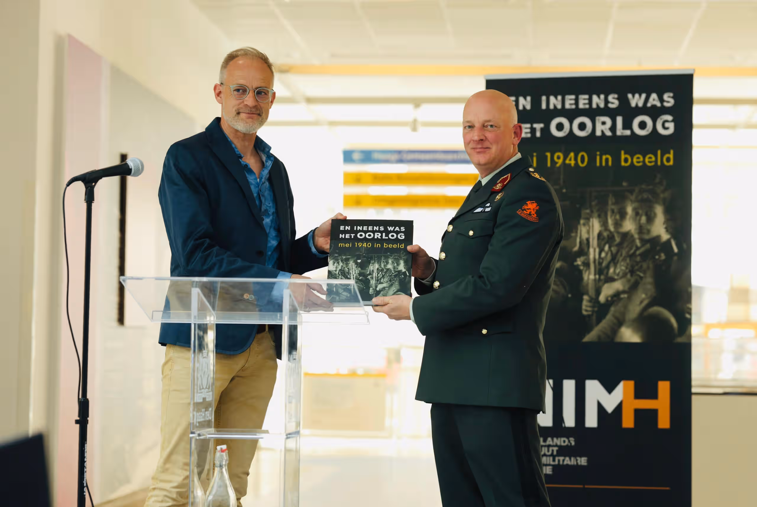 Op de foto wordt het boek door NIMH-medewerker overhandigd aan generaal-majoor Roy Sillen. Op de achtergrond is een NIMH-banner te zien.