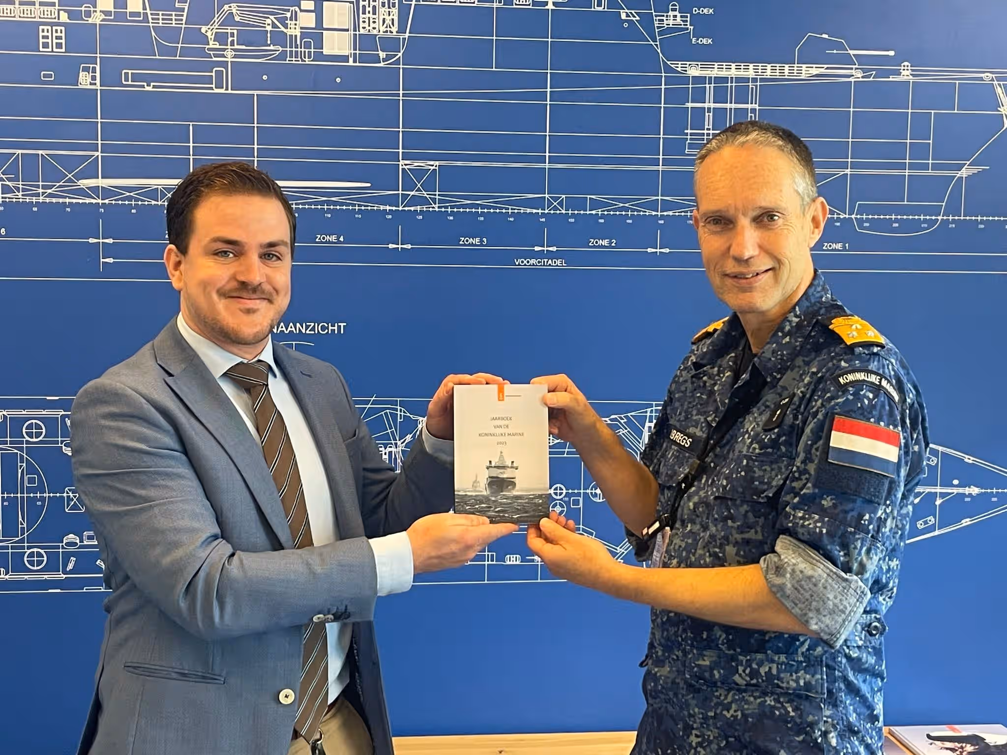 Kleurenfoto. Er staan twee mannen op de foto. De man links heeft een grijs pak aan. De man rechts heeft zijn marine-camouflage uniform aan. Samen houden zij het jaarboek vast. Op de achtergrond een muur waarop een blauwdruk van een marineschip te zien is.