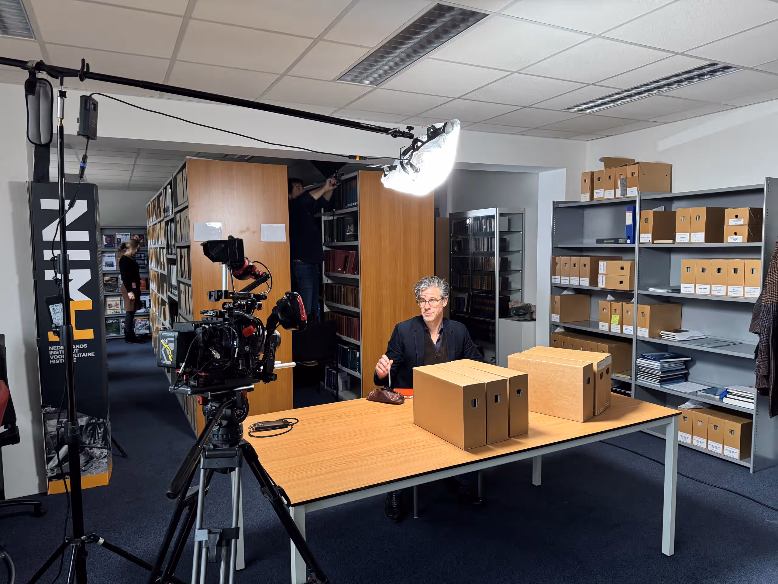 Kleurenfoto. Een man zit aan een bureau, waar allerlei archiefdozen op staan. Achter hem is een persoon bezig een lichtkap goed te stellen. Linksvoor staat de camera klaar.