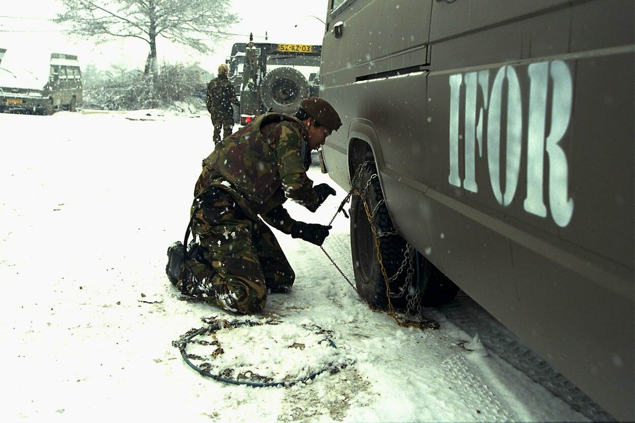 Een militair zit op zijn knieën in de sneeuw naast het wiel van een voertuig met de letters 'IFOR' op de zijkant gespoten, op de achtergrond staan nog 2 voertuigen en een persoon met rug naar de camera.