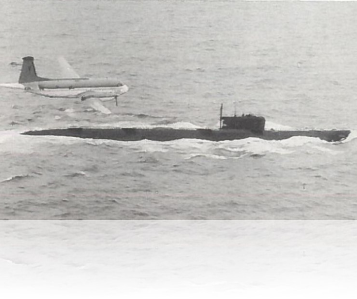 Zwart-witfoto van een onderzeeboot die boven water vaart. Boven de onderzeeboot vliegt een klein vliegtuig.
