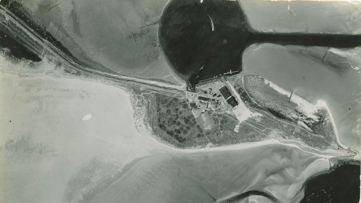 Vliegkamp De Mok van bovenaf gezien, voorafgaand aan de Meidagen van 1940. In een rond meer zijn aanmeersteigers voor de watervliegtuigen te zien. Dit is te onderscheiden doordat het water donker van kleur is. Op het land zijn een aantal gebouwen, waaronder hangars te zien.