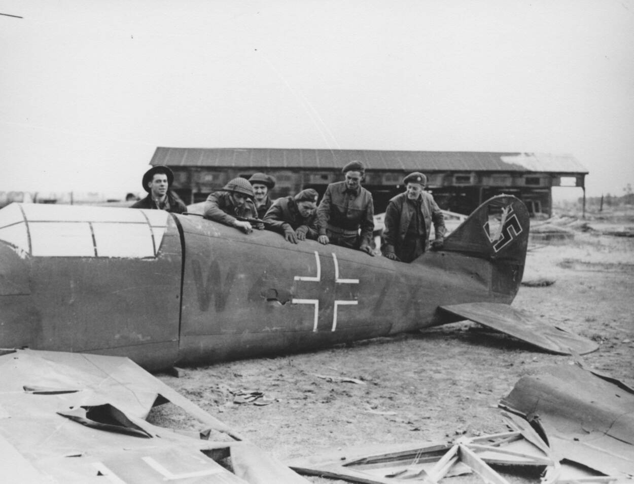 Zwart-wit foto. Zes militairen in uniformen hangen over de romp van een vliegtuig heen. Op de achtergrond is een gebouw te zien.