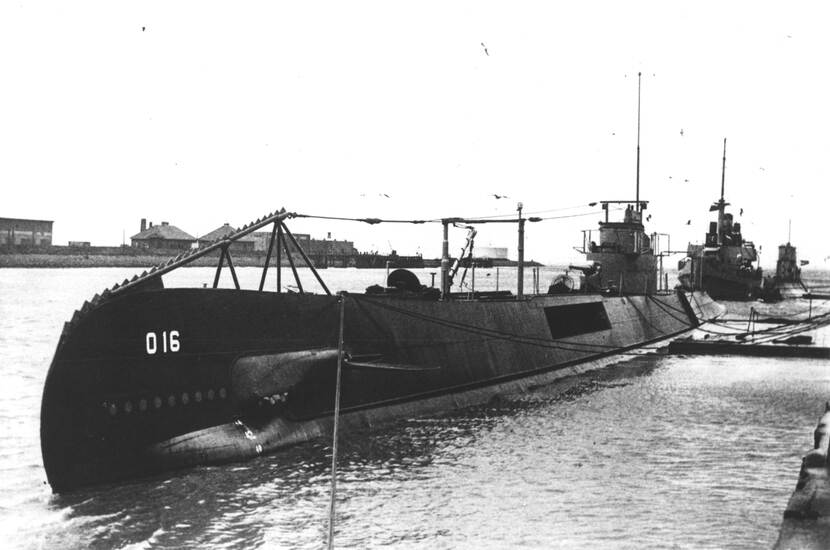 Zwart-witfoto van een aangemeerde onderzeeboot in een haven, op de zijkant staan de letters 'O 16', op de achtergrond is nog een aangemeerde onderzeeboot te zien en een schip.