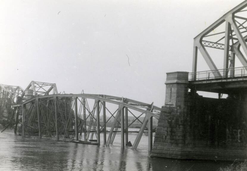 Zwart-witfoto van een ingestorte spoorbrug over een rivier. Het middelste stuk is geheel ingestort en ligt gedeeltelijk onder water.