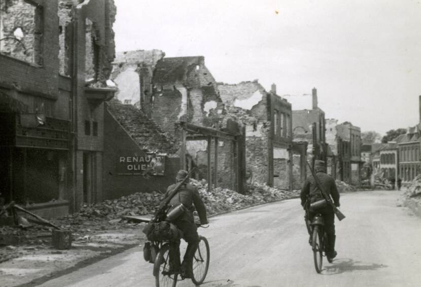 Zwart-witfoto, twee militairen fietsen door een zwaar beschadigde straat. Ze hebben geweren op hun rug. Links staan van de gebouwen in de straat enkel nog de muren overeind.