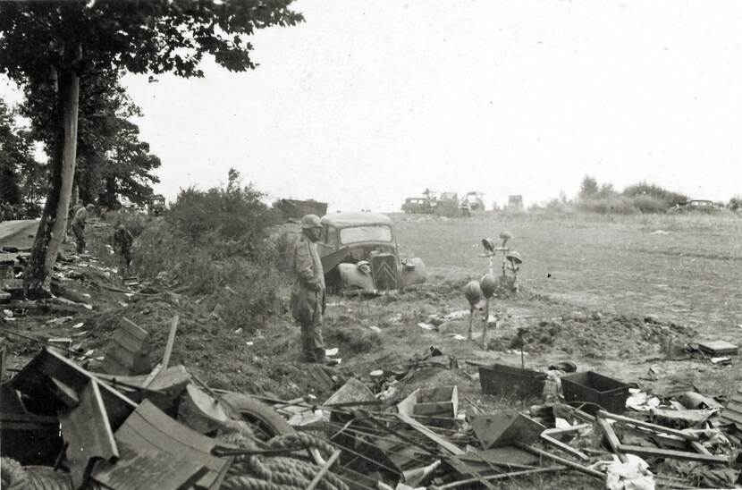 Zwart-witfoto, op de rand van een weiland staat een militair in een greppel, om hem heen ligt puin, achter hem staat in het veld een verwoeste auto. Links loopt een landweg, op de achtergrond zijn militairen in de greppel door het puin aan het zoeken.