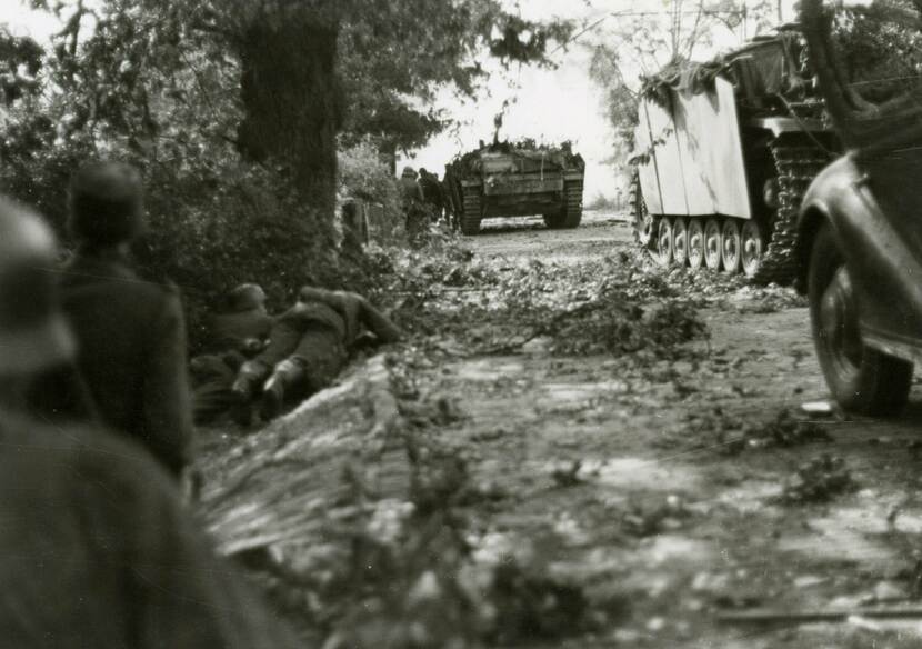 Zwart-witfoto genomen vanaf de grond, rechts een straat bezaaid met takken waar twee tanks en een auto rijden, links in de berm liggen twee militairen, links op de voorgrond twee militairen met de rug naar de camera.