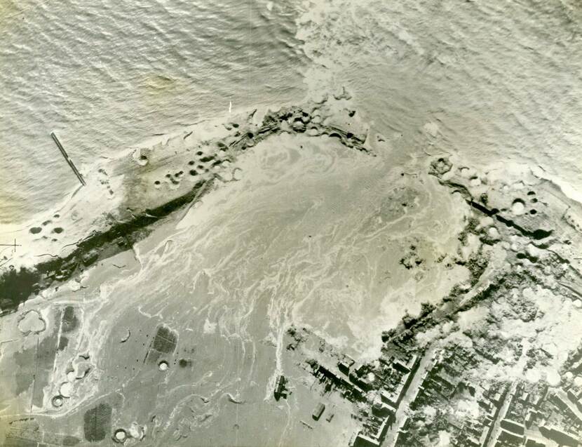 Luchtfoto in zwart-wit, in de zeedijk zijn veel bomkraters te zien, door een groot gat stroomt zeewater naar binnen, rechts een woonwijk die bijna wordt overstroomd door het binnenkomende zeewater.