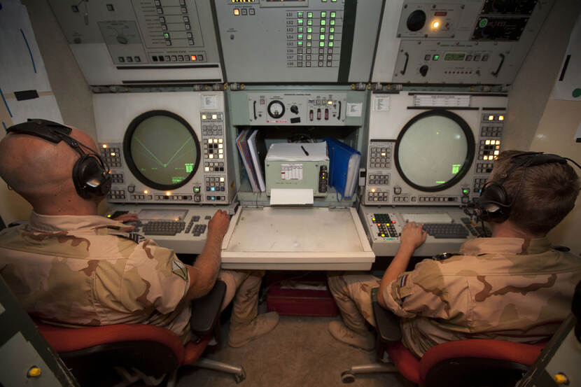 Kleurenfoto, twee militairen in camouflagekleding zitten achter radarpanelen, met vele knoppen en twee grote ronde radarschermen. De militairen hebben allebei een koptelefoon op. Tussen hen in liggen een paar mappen papier.