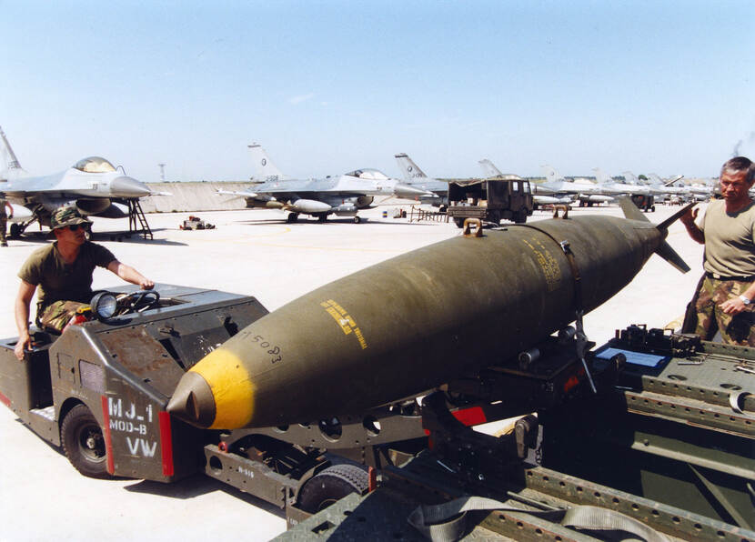 Kleurenfoto, buiten op een vliegveld. Links een militair die een kleine takelwagen bedient, de takelwagen tilt een grote bom op, rechts van de bom een andere militair. In de achtergrond staat een lange rij F-16’s. De zon schijnt, er is geen wolkje aan de lucht.
