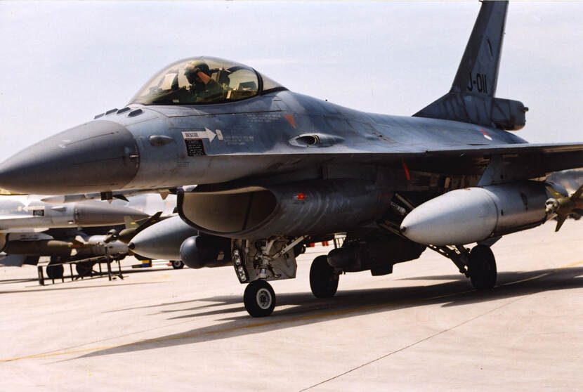 Kleurenfoto, buiten op een vliegveld. Een F-16 met onder elke vleugel een grote bom, taxiet richting de startbaan. In de cockpit is de piloot te zien, hij groet of zwaait. Op de staart van het toestel staat J-011, daaronder is de rondel van de Koninklijke Luchtmacht te zien. Links in de achtergrond een andere F-16.