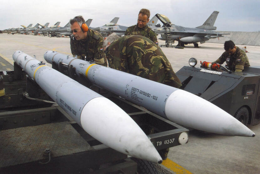 Kleurenfoto, buiten op een vliegveld. Twee militairen in camouflagekleding zijn met twee lange raketten in de weer, een derde (ook in camouflagekleding) staat ernaast en kijkt toe. Rechts van hen een vierde militair die een kleine takelwagen bedient. De meest linker militair draagt een oranje koptelefoon en een zonnebril. De militair op de voorgrond buigt voorover en verdwijnt half achter één van de raketten. In de achtergrond staat een lange rij F-16’s.