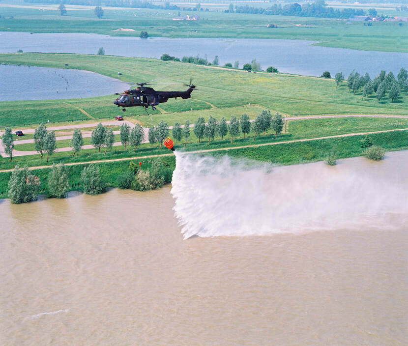 Kleurenfoto. Een helikopter vliegt boven water, daaronder hangt een oranje bak aan een kabel. Uit de bak komt water dat in een grote hoeveelheid in het water daaronder terechtkomt. Achter de helikopter een uiterwaard met bomen en gras, daarachter nog meer water, afgewisseld met andere uiterwaarden.