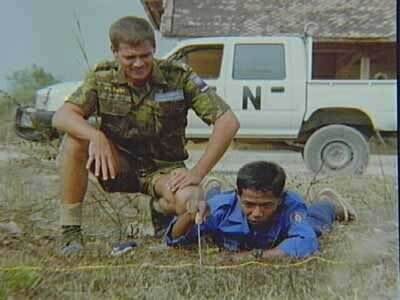 Kleurenfoto, een militair in camouflagekleding zit op zijn hurken naast een Cambodjaan die op zijn buik op de grond ligt, de Cambodjaan prikt in de grond, achter hen een witte VN-wagen.