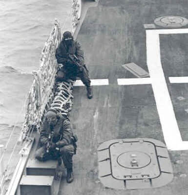 Zwart-witfoto, twee militairen geheel in zwart gekleed en met bivakmutsen zitten met geweren in de aanslag langs een hek langs de rand van het schip. Rechts van hen zijn witte strepen aan boord, links is zeewater.