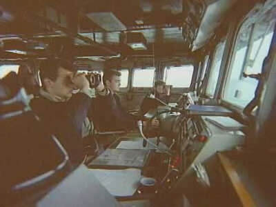 Kleurenfoto, drie mannen in de navigatiekamer van een marineschip. De middelste houdt het roer vast, terwijl de andere twee met een verrekijker recht vooruit naar buiten kijken. Er zijn ramen voor en naast hen, zij zitten achter meerdere bestuurstafels.