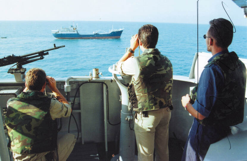 Kleurenfoto, drie mannen vanaf de rug gezien kijken aan boord van een marineschip naar een schip verderop op zee. Twee kijken door een verrekijker, de derde draagt een zonnebril. Links op de reling staat een mitrailleur.