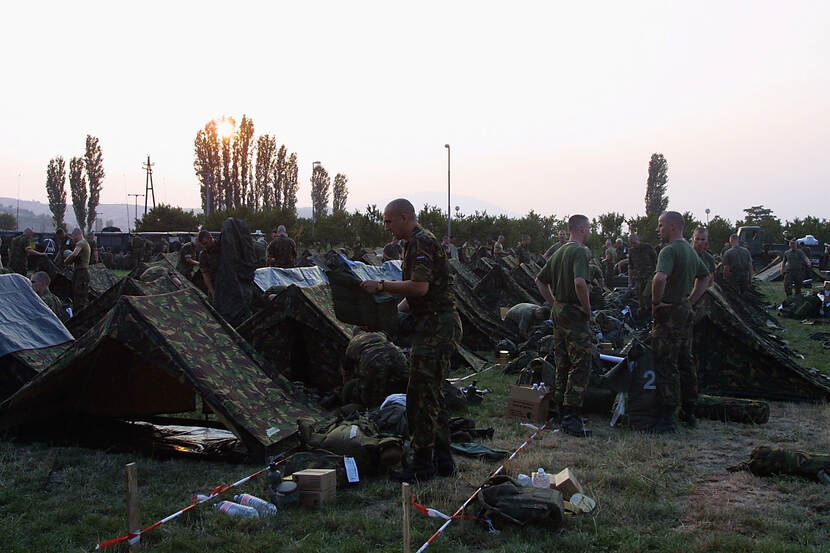 Kleurenfoto, buiten. In een grasveld staan tientallen kleine tentjes opgesteld, allemaal in camouflagekleuren. Tussen de tenten staan, zitten en lopen militairen, vaak zijn ze met elkaar in gesprek. In de voorgrond op de grond twee paaltjes met aan elk een rood-wit lint die door het gras strekt. In de achtergrond bomen, waar de ondergaande zon doorheen schijnt.
