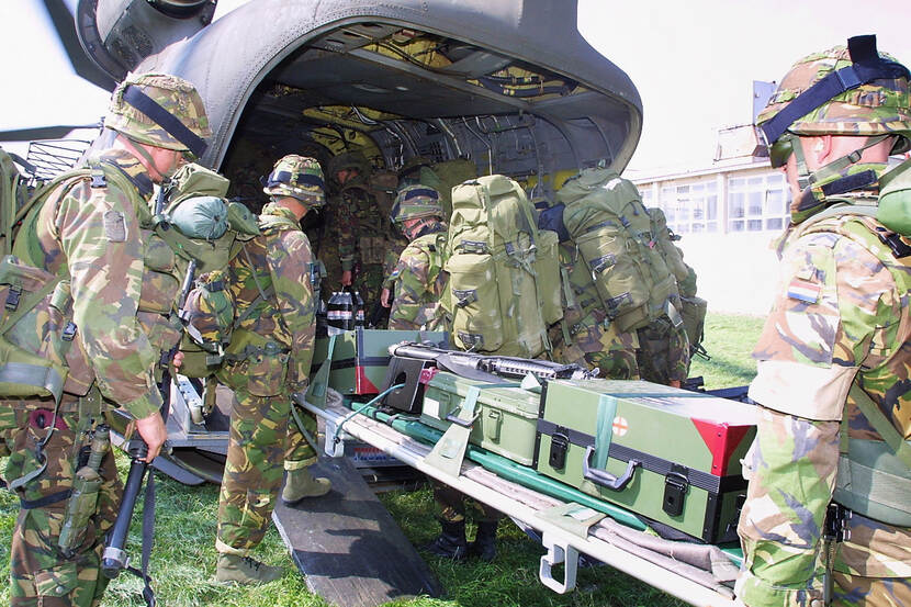 Kleurenfoto, buiten. Vijf militairen staan achter een transporthelikopter, zij zijn in vol ornaat, inclusief grote rugtassen en helm. De laadklep van de helikopter is open en de militairen tillen een brancard naar binnen. Op de brancard liggen kisten, sommigen daarvan zijn voorzien van een wit vlakje met een rood kruis.