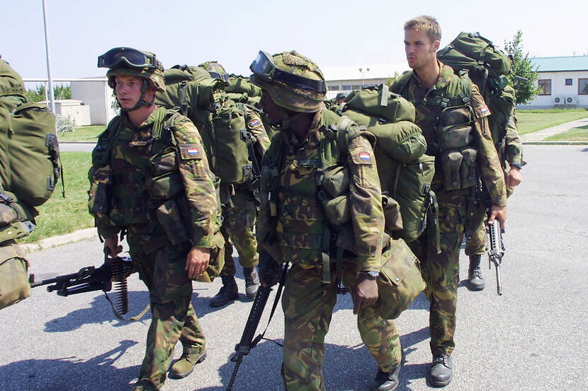 Kleurenfoto, buiten. Een groep van zes militairen loopt over straat, zij dragen allemaal camouflagekleding en zijn bepakt met grote rugtassen, helmen en wapens. De rechter militair heeft zijn helm af, een andere militair draagt een mitrailleur nonchalant in zijn rechterhand. In de achtergrond lage gebouwen en een lichtmast.