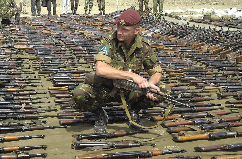 Kleurenfoto, buiten. Een militair in camouflagekleding, met opgestroopte mouwen en een rode baret zit op z’n hurken, hij houdt een geweer op zijn schoot. Om hem heen liggen honderden geweren op de grond, allemaal keurig in rijen naast elkaar. In de achtergrond aan de rand van de verzameling geweren, zijn de benen van andere militairen te zien.