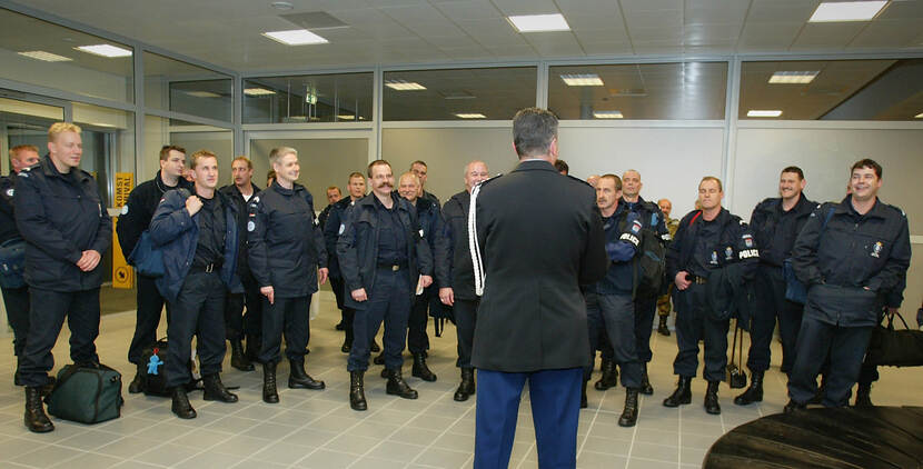 Kleurenfoto, binnen. Een groep van ongeveer zeventien militairen staan in een hal op een rij, zij dragen allemaal donkerblauwe jassen en kleding en zwarte kisten. Voor hen staat iemand die hen toespreekt, hij staat met zijn rug naar de camera, om zijn linkerschouder hangt een wit koord.