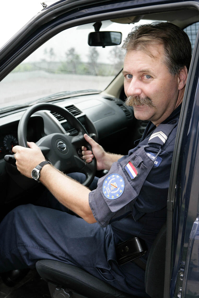 Kleurenfoto, een man met snor, gekleed in een blauw uniform, zit in een zwarte auto achter het stuur.