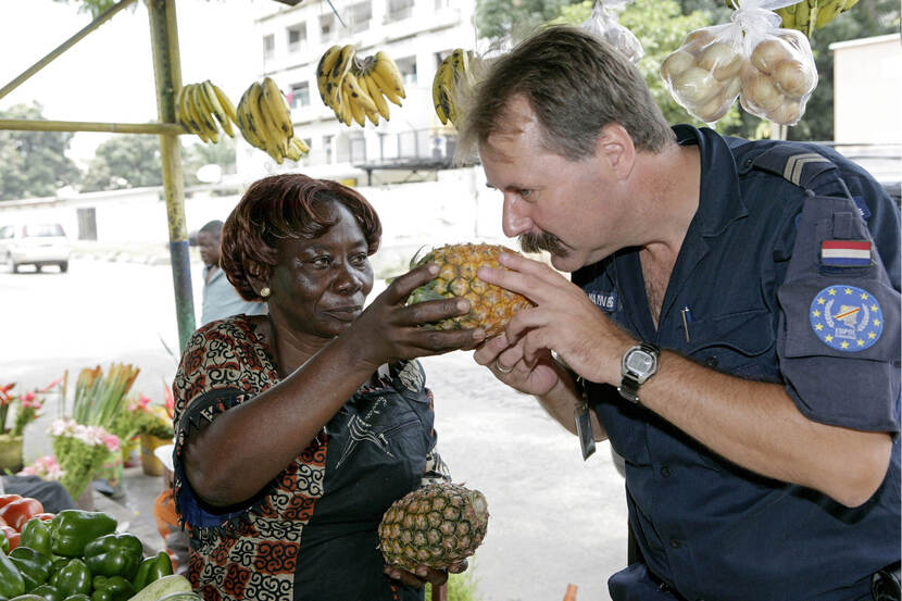 Kleurenfoto, rechts een man met snor, gekleed in een blauw uniform, links een marktvrouw. Ze houden samen een ananas vast, terwijl de man eraan ruikt. Om hen heen liggen bloemen en fruit.