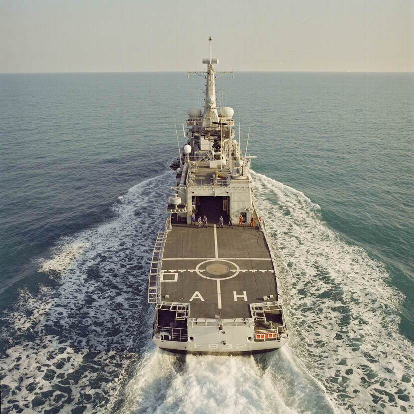 Kleurenfoto, van boven genomen. Een marineschip vaart op zee, van achteren gezien. Naast en achter het schip schuimt het water wit op, de rest van de zee is blauw, de lucht boven de horizon grijs. Op het dek staan zes personen naar de camera te kijken. Op het achterdek staan de letters A en H geschilderd.
