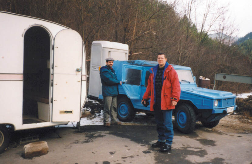 Kleurenfoto, buiten. Twee mannen staan voor een geheel blauwe jeep, één van hen staat houdt de openstaande bijrijdersdeur vast, ze kijken allebei naar de camera. Achter de jeep een mobiel toilet, links een caravan met open deur, onder de deur een steen die als opstapje gebruikt wordt. Meteen achter het toilet en de caravan een bos.